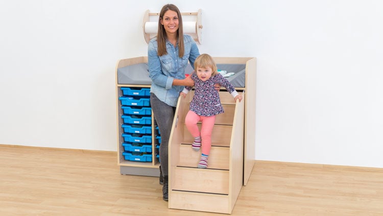 Kind klettert mit Hilfe einer Erzieherin von einem Wickeltisch mit Treppe aus dem Betzold Online-Shop.
