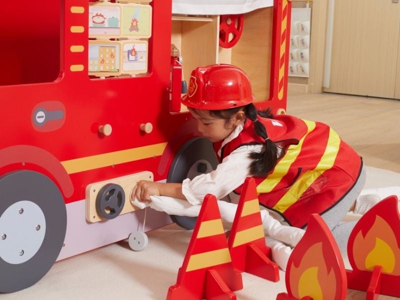 Mädchen in Feuerwehr-Kleidung schließt einen Schlauch an ein Spielmöbel-Feuerwehrauto an