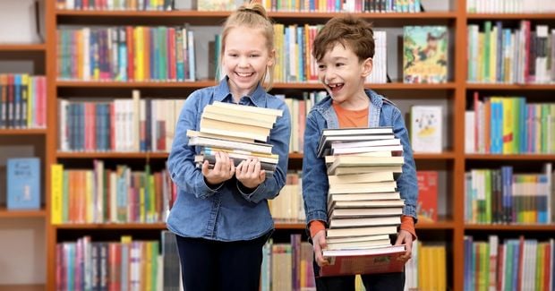 Morgenkreis Lesen Zwei Kinder stehen in einer Bibliothek und tragen jeweils einen Stapel Bücher